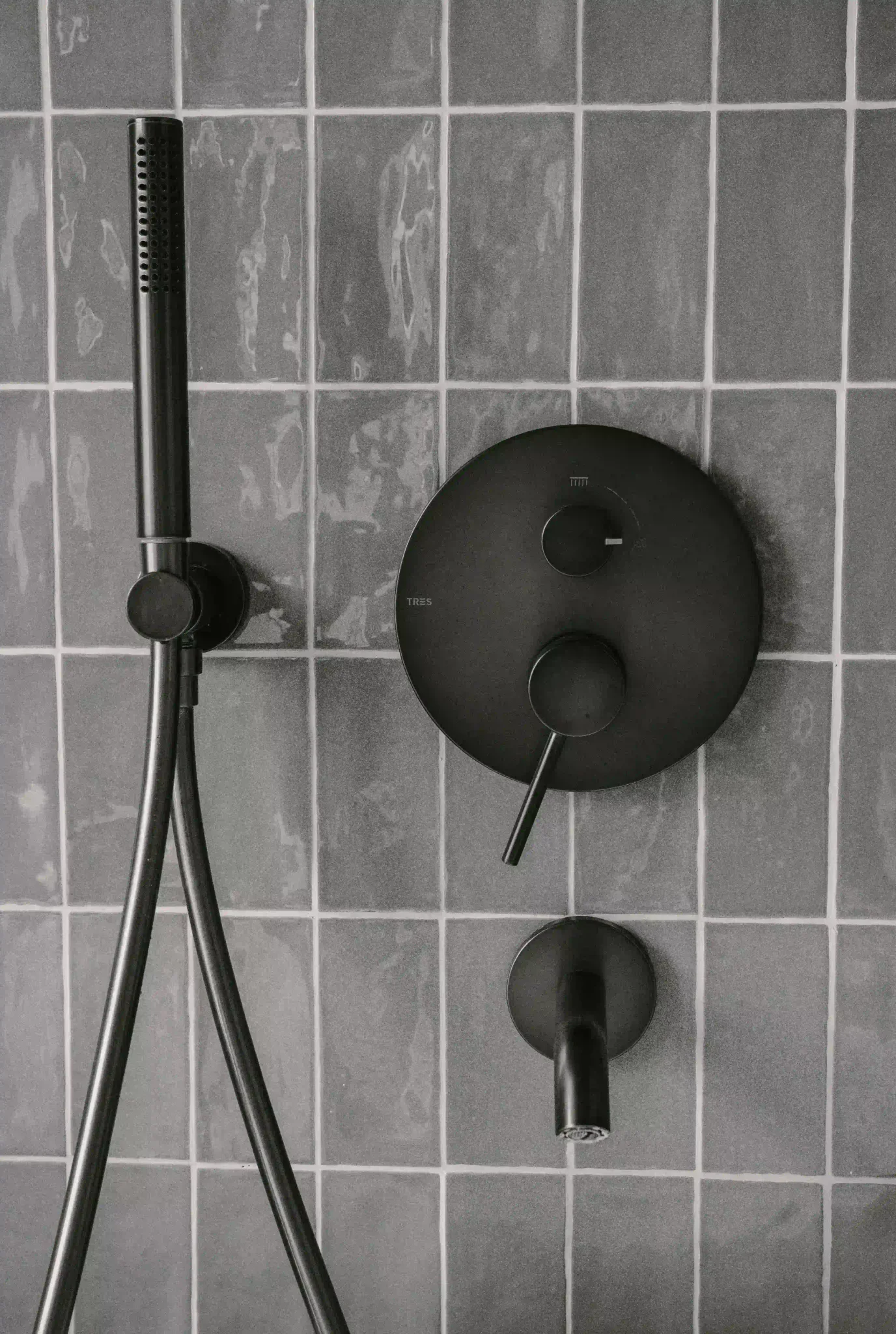 Matte black shower mixer and handset mounted on textured gray square wall tiles — detail of finished tile work